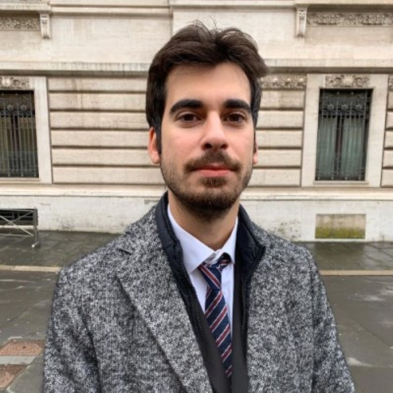 Photo of Roberto. He is standing in front of a stone building, wearing a grey coat, white shirt and stripey tie. he has dark hair and a beard